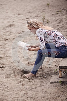 One young girl, sitting on steps on beach sand, summer, happy smiling, reading a book outdoors. side view