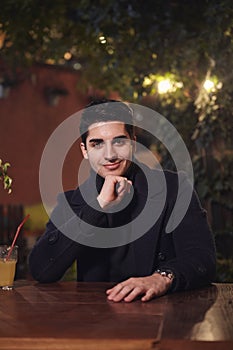 One young handsome man portrait, sitting in cafe garden at table, night outdoors, looking to camera. upper body shot