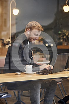 One young happy man, sitting in coffee shop and using his laptop, typing on keyboard, looking to computer screen. Shoot thought