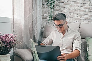 One young happy man surfing the net using his laptop at home sitting on the sofa enjoying free time and relaxing alone.