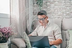 One young happy man surfing the net using his laptop at home sitting on the sofa enjoying free time and relaxing alone.