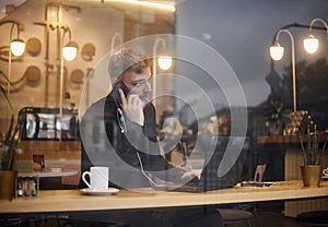 One young man, 20-29 years, sitting in coffee shop and using, his laptop while talking over phone. Shoot thought window outside