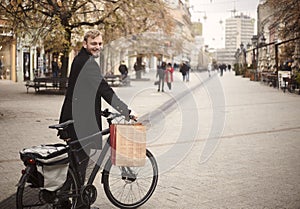 One young man, 20-29 years, standing in, pedestrian only, street holding his bicycle, and smiling to camera, with shopping bag.