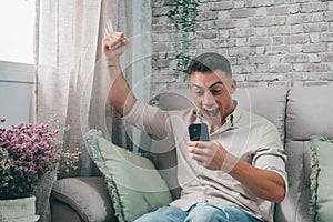 One young man celebrating some winning on his phone at home. Euphoric teenager having fun watching football or investment on