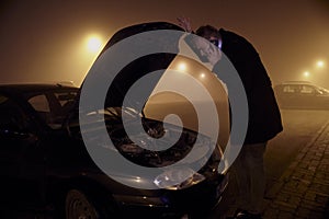 One young man, at dark night, looking at car engine, with car hood open. rural area