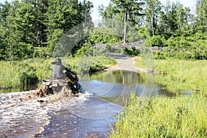 Man driving motocross ATV quad through splashing river lake water. Foy, Sudbury, Ontario, Canada.