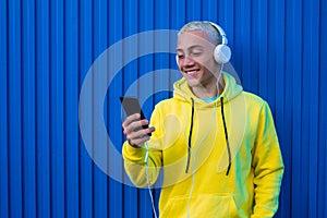 One young man hearing and listening music smiling and looking at the phone having fun - blue colorful wall at the background -