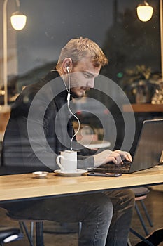 One young man, sitting in coffee shop and using his laptop while typing on keyboard. Shoot thought window outside with