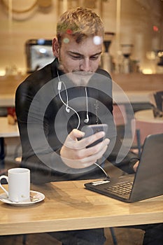 One young man, sitting in coffee shop and using his smart-phone, with laptop and cup nearby. Shoot thought window outside with