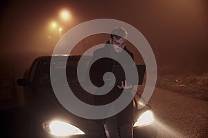 One young man using his laptop computer, on a car hood. It`s dark and night time, in rural area