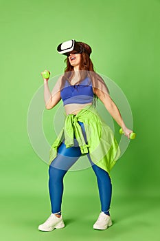 One young pretty girl, woman wearing vr glasses doing workout with dumbbells and smiling at camera over green background