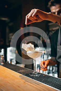 One young professional bartender decorating alcohol cocktail with flakes or dried flowers standing at bar counter