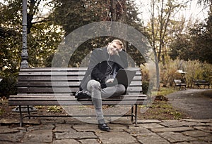 One young relaxed man, sitting on bench in public park, using laptop,
