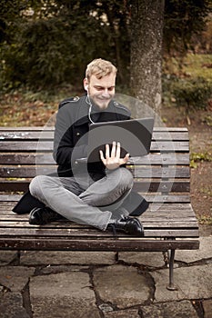 One young relaxed and smiling man, sitting casually on bench with crossed legs in public park,