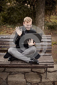 One young relaxed and smiling man, sitting casually on bench with crossed legs in public park, using laptop with video call