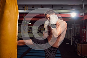 one young shirtless man, boxer leg hitting punching bag, practicing indoors gym room, wearing boxing gloves, upper body shot.