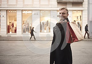 One young smiling and happy man, 20-29 years old, handsome and stylish, looking to camera while holding two shopping bags on his