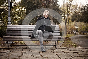 One young smiling and happy man, sitting on bench in public park, using laptop,