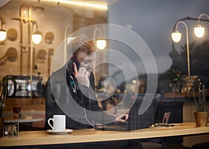 One young smiling man, 20-29 years, sitting in coffee shop and using, his laptop while talking over phone. Shoot thought window