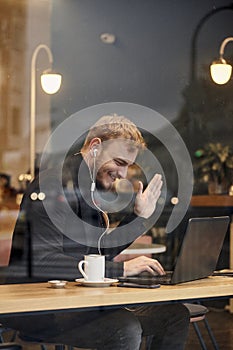 One young smiling man, sitting in coffee shop and using his laptop, gesturing with his hand Hello or Hi, waving his hand over