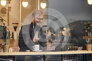 One young smiling man, sitting in coffee shop and using his laptop, gesturing with his hand peace sign with two fingers over
