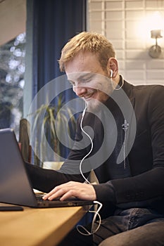 One young smiling man, sitting indoors in coffee shop and using his laptop, earbuds in ear