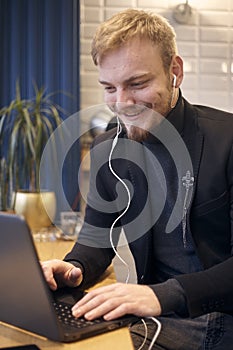 One young smiling man, sitting indoors in coffee shop and using his laptop, earbuds in ear