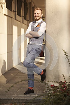 One young smiling man, 20-29 years old, wearing hipster suit, standing, leaning to building column, looking to camera