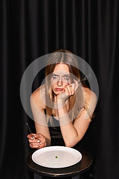 One young thin girl, fashion model sitting at table and starting eating almost empty plate over black background