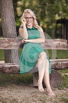 One young woman, 25 years old, sitting in wood bench in park, green dress, looking to camera. full length shot, barefoot