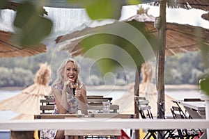 One young woman, 25 years old, smiling sitting in beach cafe outdoors, sunshades leaves. summer vibes