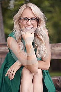 One young woman eyeglasses portrait, sitting in wood bench, green dress, 25 years old, smiling happy, looking to camera.