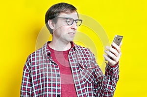 Onfident smiling guy in red checkered shirt and glasses looking at smartphone on yellow background