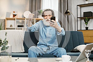 onfident stylish man with grey beard smiling happily sitting on the couch at cozy living room