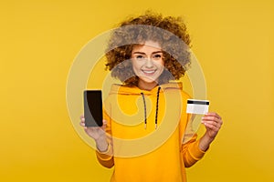 Online banking app, electronic money. Portrait of happy curly-haired woman in urban style hoodie showing credit card