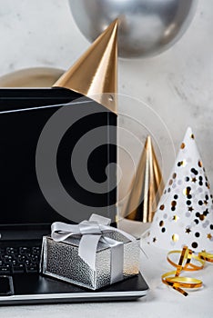 Online birthday celebration concept. Silver festive box on laptop with party hats and balloons