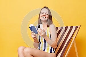 Online booking. Traveling abroad. Satisfied young woman wearing striped swimming suit sitting on deck chair using cell phone,