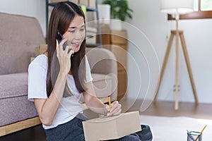 Online business concept, Asian business woman talks on smartphone and writes address on parcel box