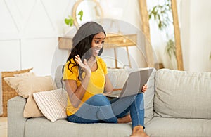 Online communication. Young black woman sitting on sofa at home and talking to family, friends or coleagues on laptop photo