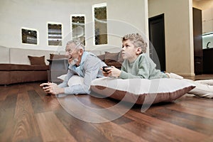 Online competiotion. Side view of a grandfather with grandson lying on pillows on the floor and playing video games