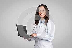 Online consultation service. Confident woman doctor using laptop, standing on grey studio background smiling at camera