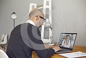 Online doctor. Senior man talking to a doctor using a video call laptop video camera sitting at a table at home.