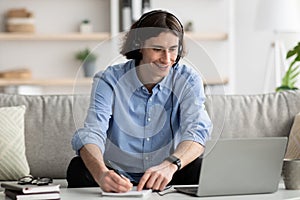 Online education concept. Happy young guy in headset studying with laptop at home, taking notes while watching webinar