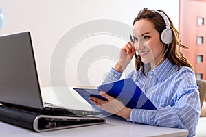 Online education concept, woman in blue shirt with headphones and notebook in hands sitting at laptop