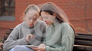 Online education and knowledge for kids. Girls students sitting in park on bench with a modern digital tablet. Teens