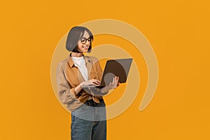 Online education programs. Excited young lady holding and using newest laptop over yellow background, free space
