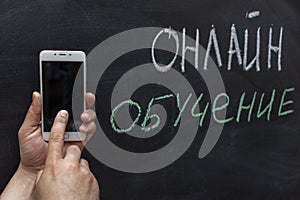 Online education in Russian text in white and green chalk on the chalk Board. Hand with a mobile phone. The concept of online