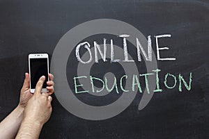 Online education text in white and green chalk on the chalk Board. Hand with a mobile phone. The concept of online education