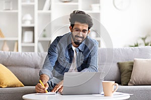 Online Education. Young indian man using laptop and taking notes at home