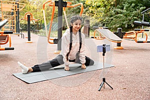 Online fitness training in modern outdoors sportground performed by young Indian woman doing twine and looking at smartphone